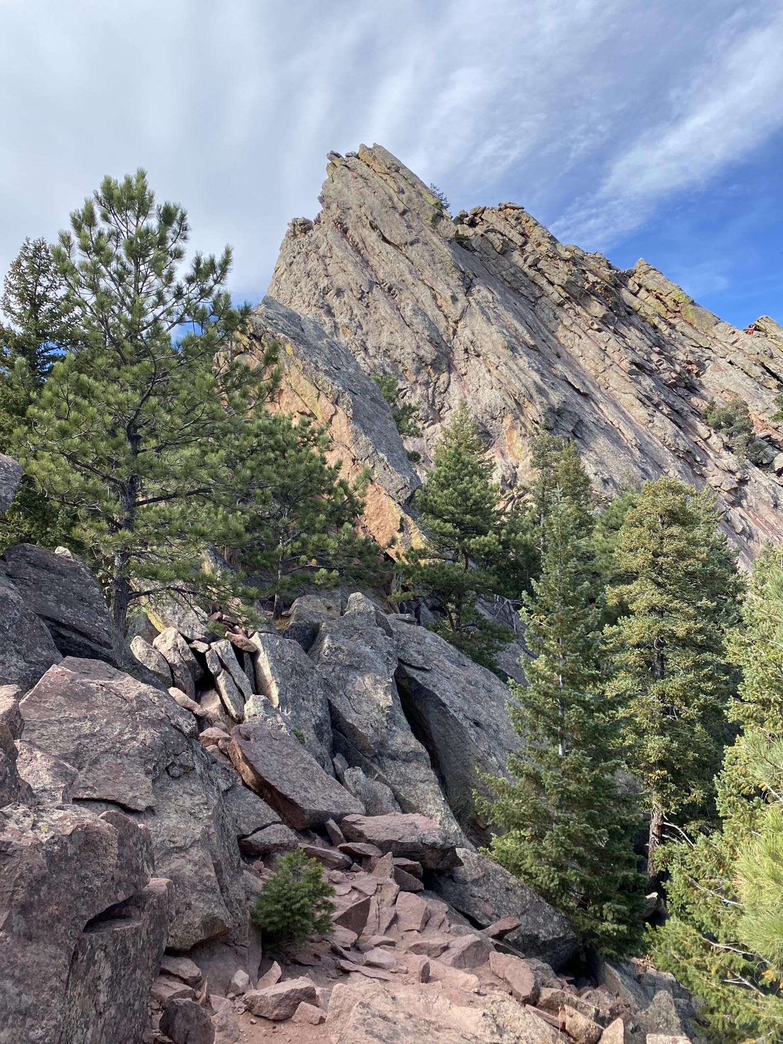Steep, jagged rock formation surrounded by pine trees and boulders under a cloudy sky.