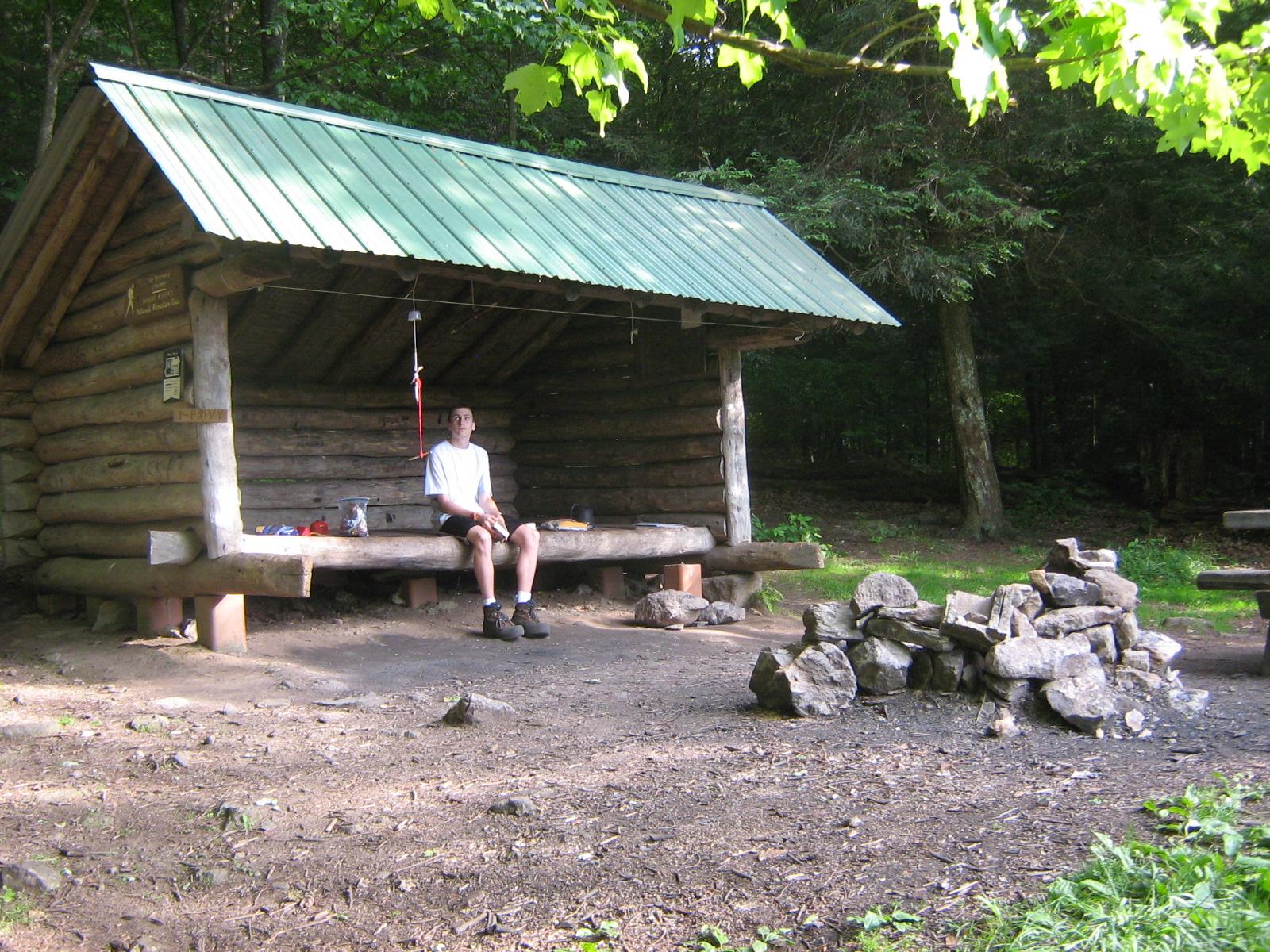 Person resting inside a rustic log shelter with a green roof in dappled sunlight.