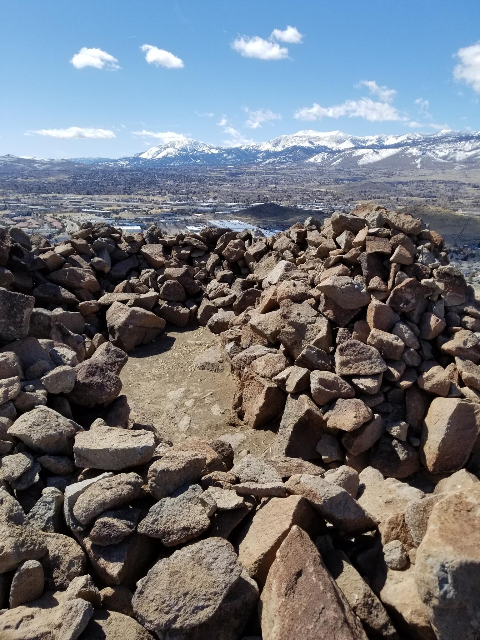 Rocky circular structure on a hilltop overlooking a valley and snowy mountains.