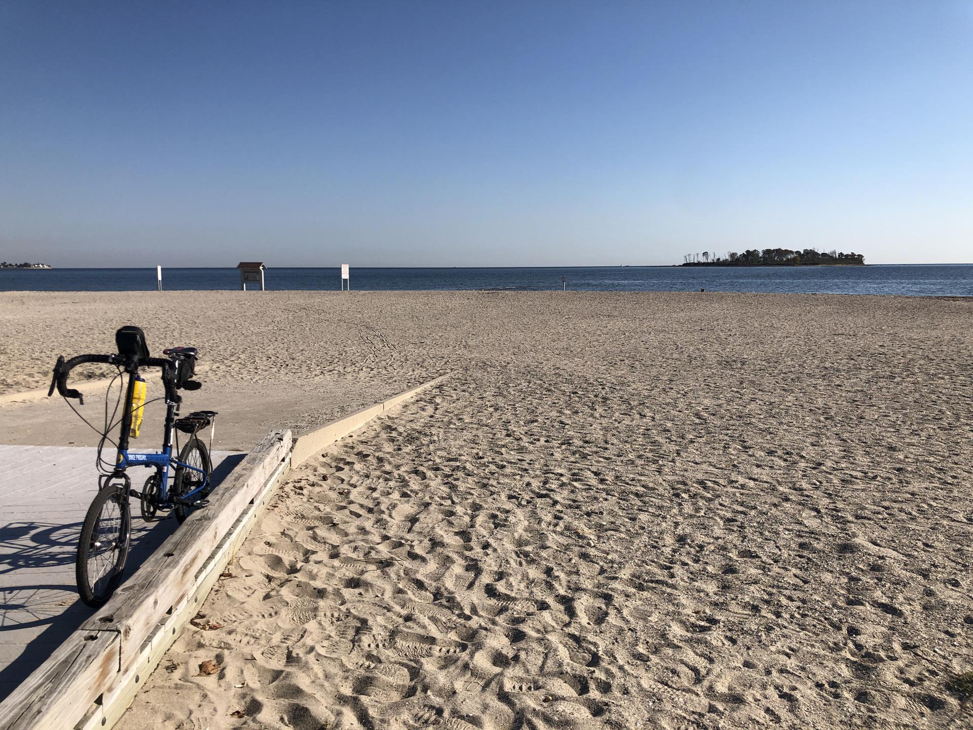 A blue folding bicycle parked on a boardwalk next to a wide sandy beach.