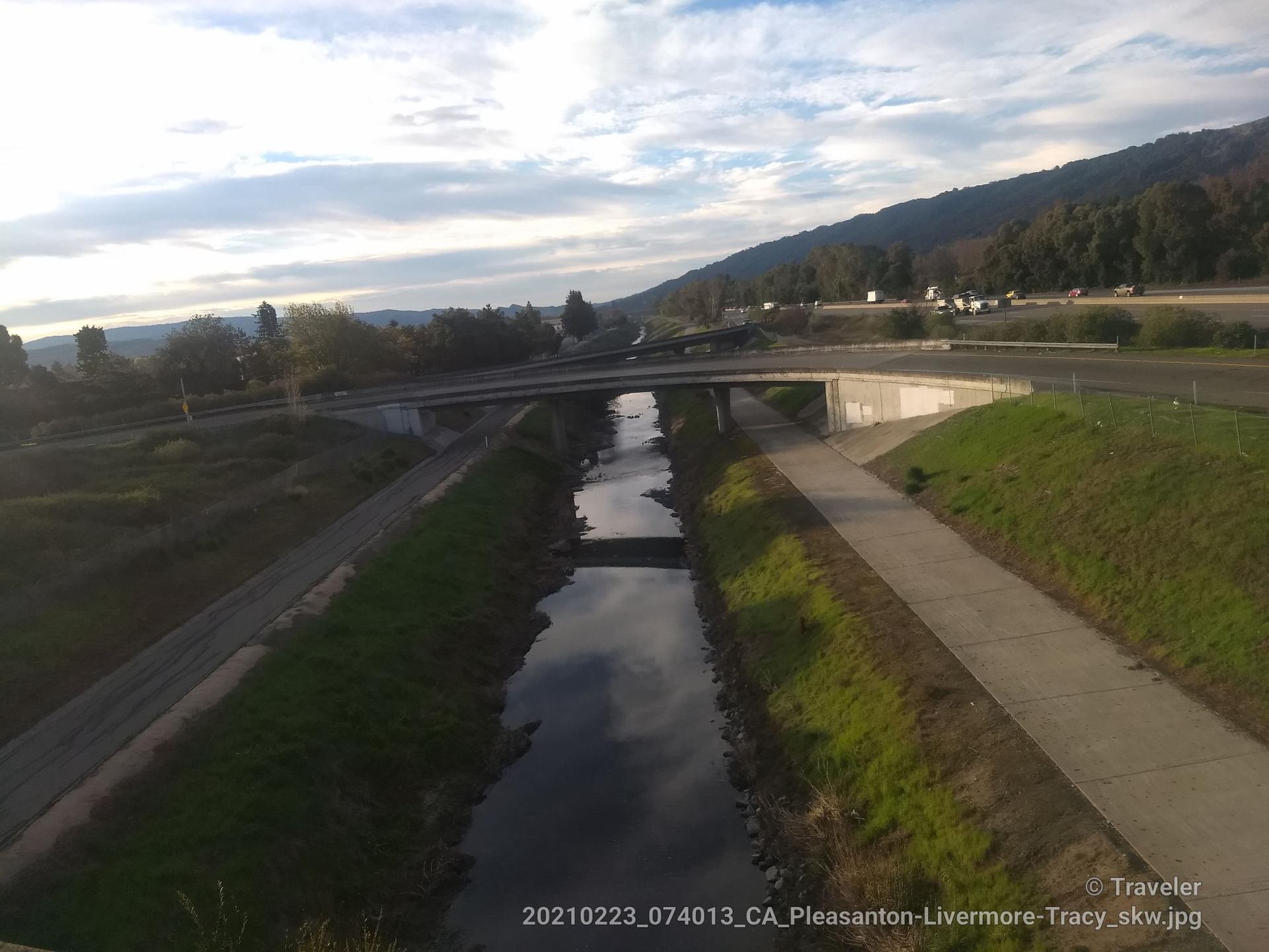 A calm river flows under a highway bridge with green hills.
