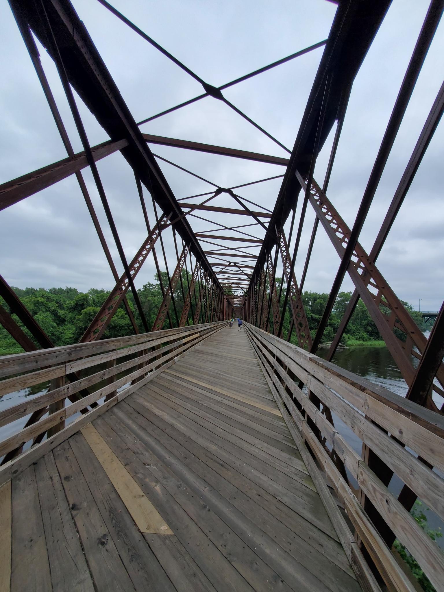 Long wooden and metal truss bridge over river under overcast sky, people walking.