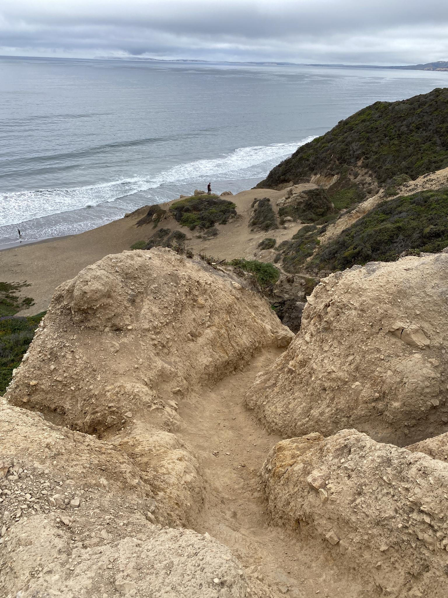 Sandy path on a cliff overlooking a beach and ocean under an overcast sky.