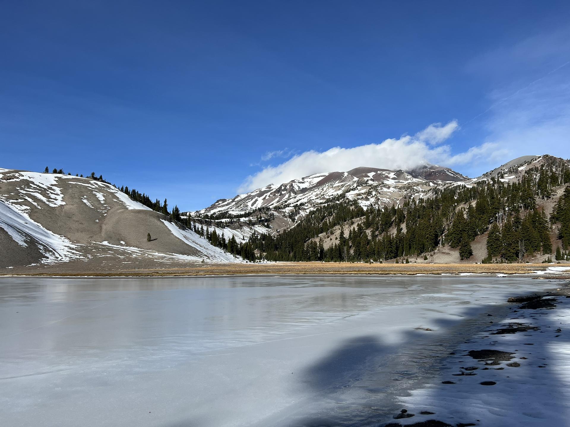 Frozen lake reflects bright blue sky, surrounded by snowy mountains and pine trees.