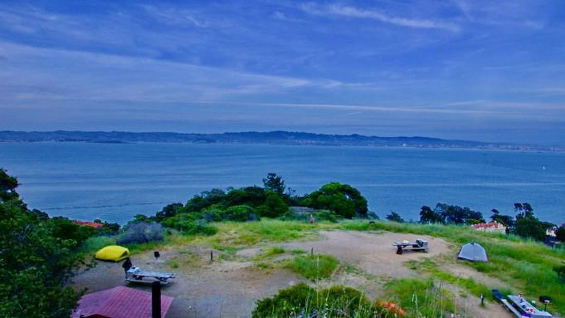 Overcast view of a wide bay, distant hills, and a hillside campsite.