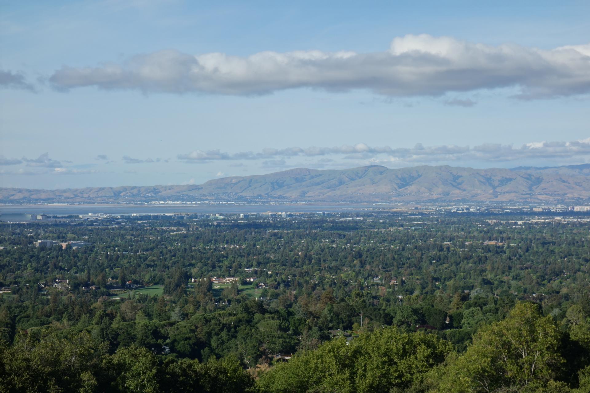 Panoramic view of a green valley, distant city, and mountains under clouds.