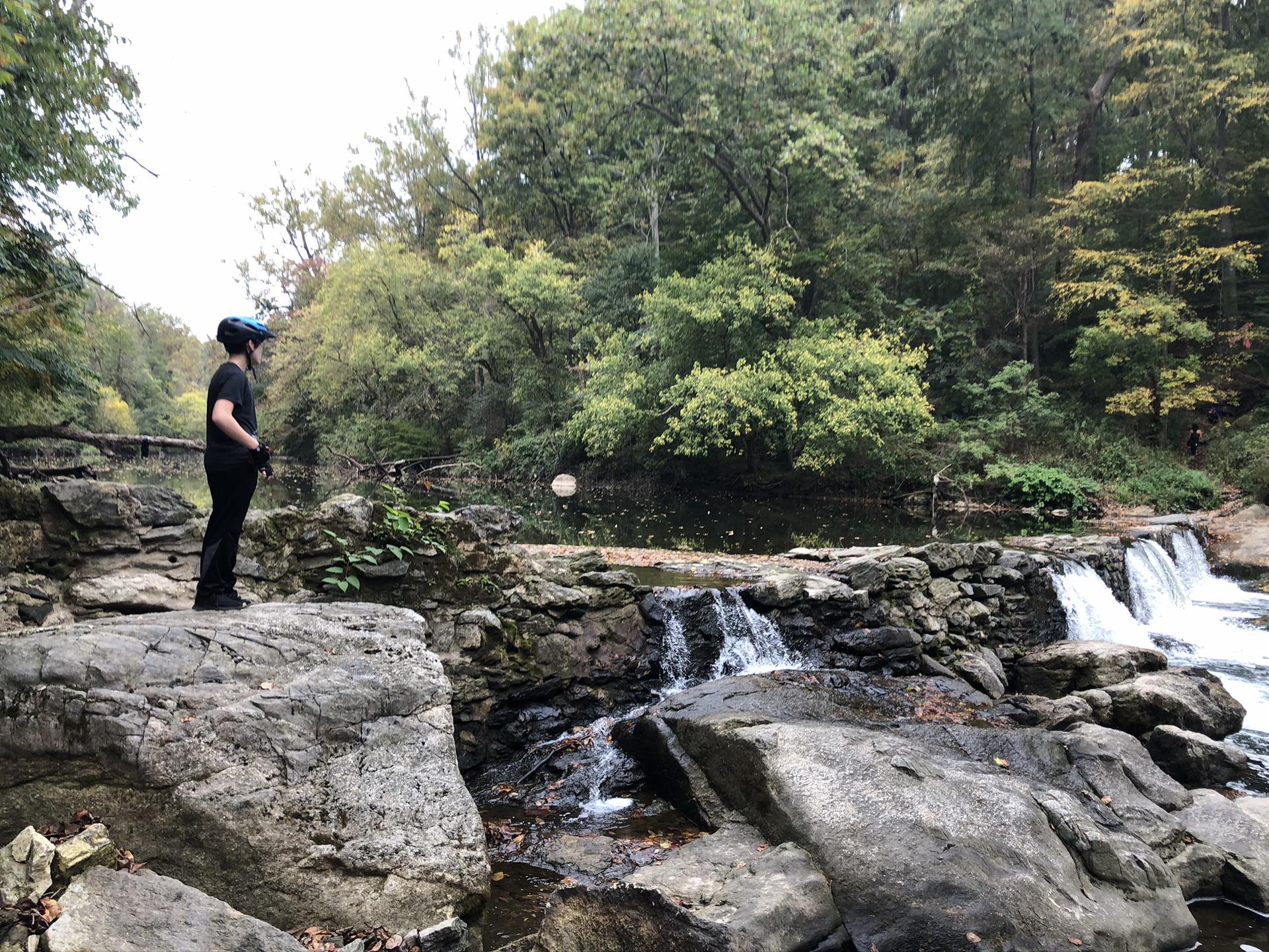 A person stands on large rocks, observing a river with small waterfalls.