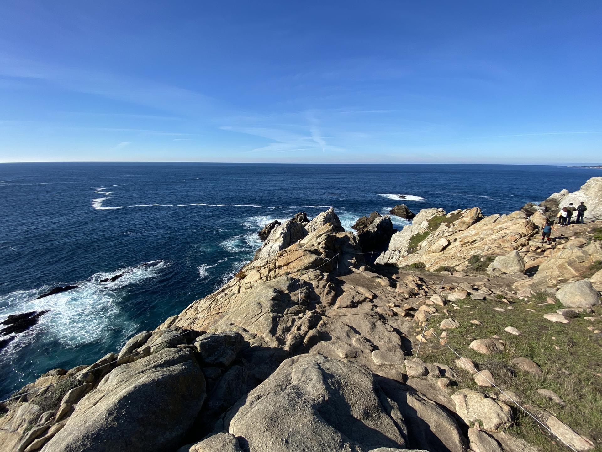 Sunny coastal view with rocky cliffs, deep blue ocean, and distant people.