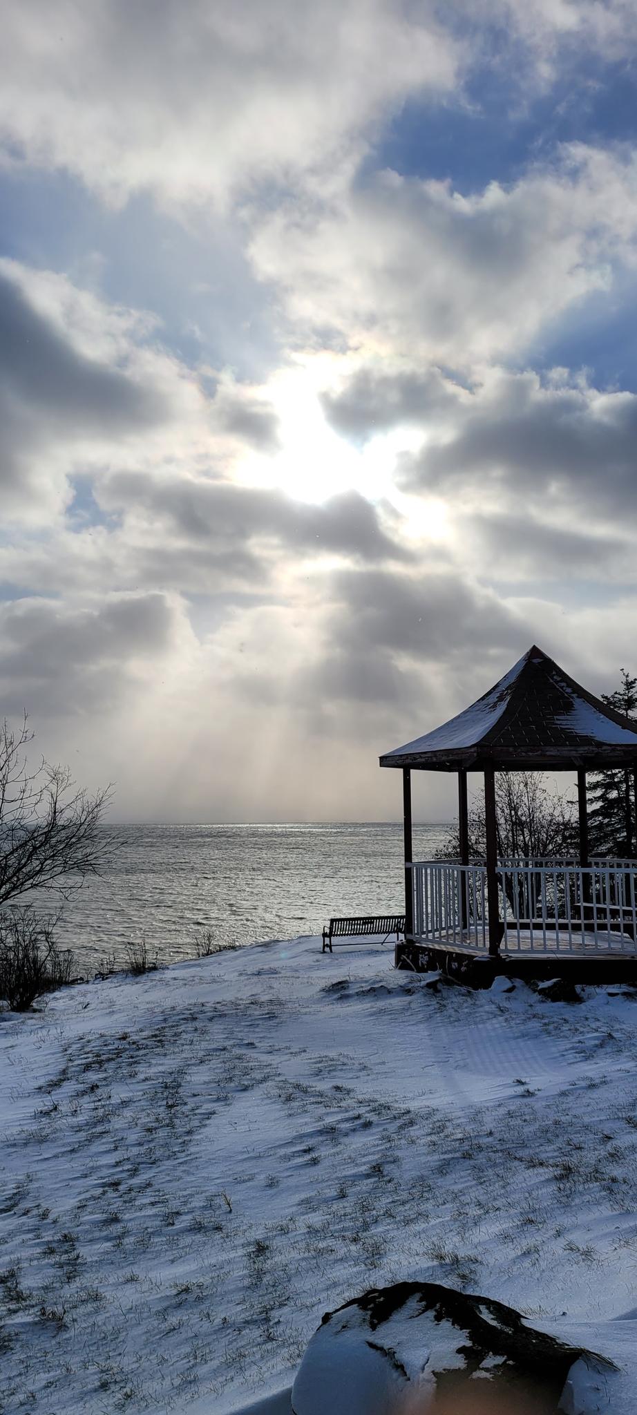 Snowy landscape with a gazebo overlooking water under dramatic, sunlit clouds.
