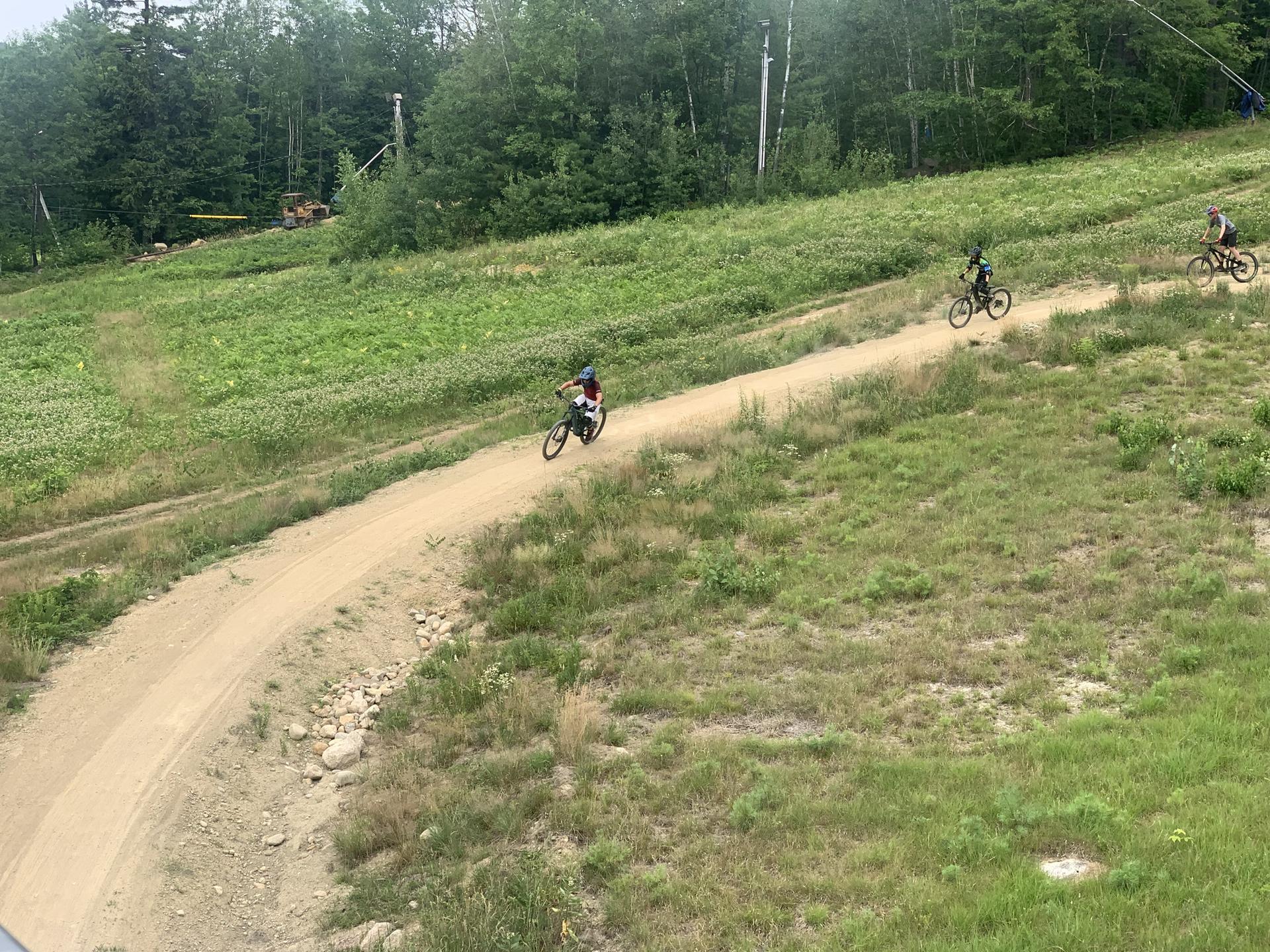 Three mountain bikers descend a wide dirt path on a grassy hill.