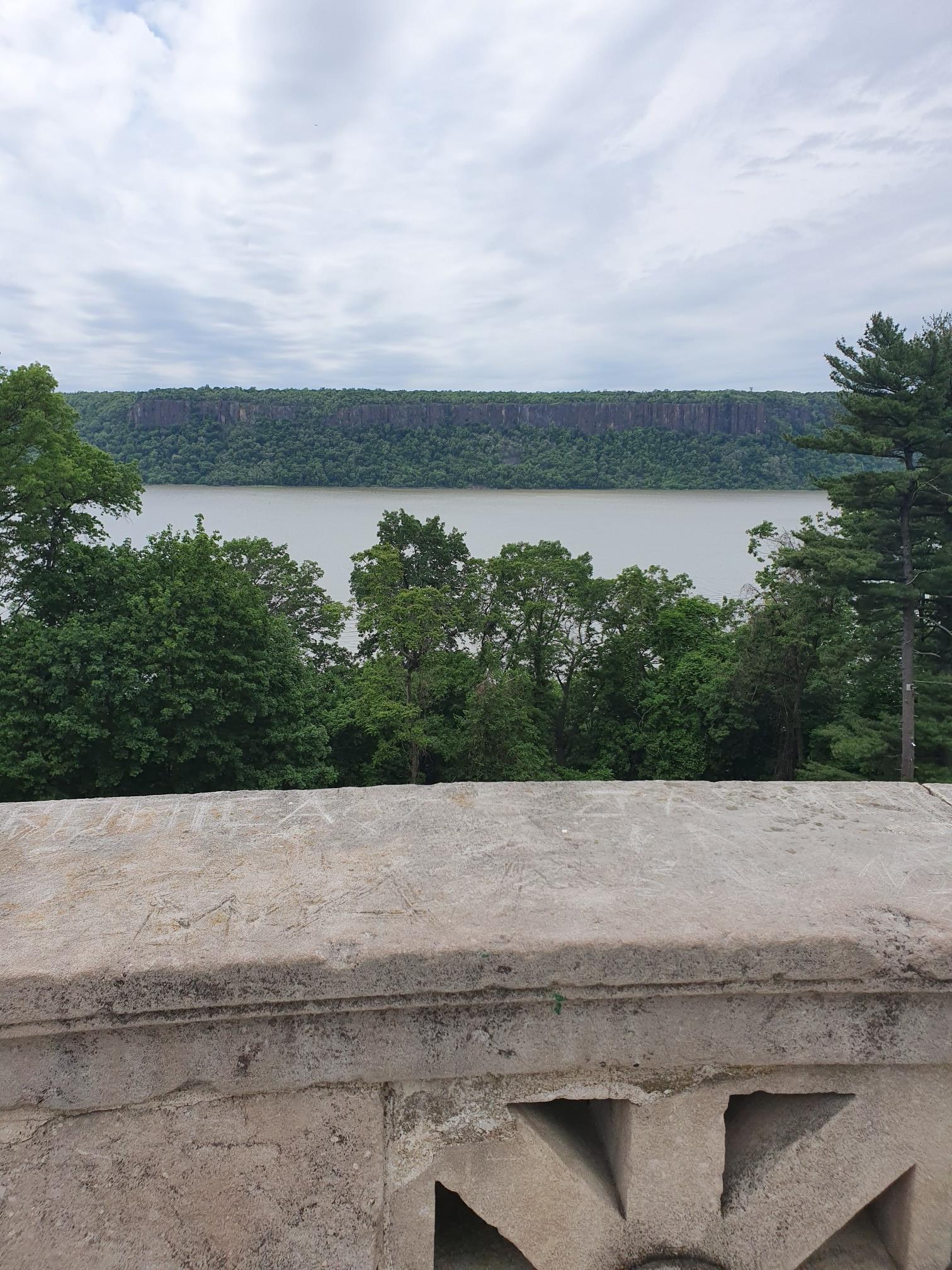 Stone railing overlooking a wide river, distant forested cliff, cloudy sky.