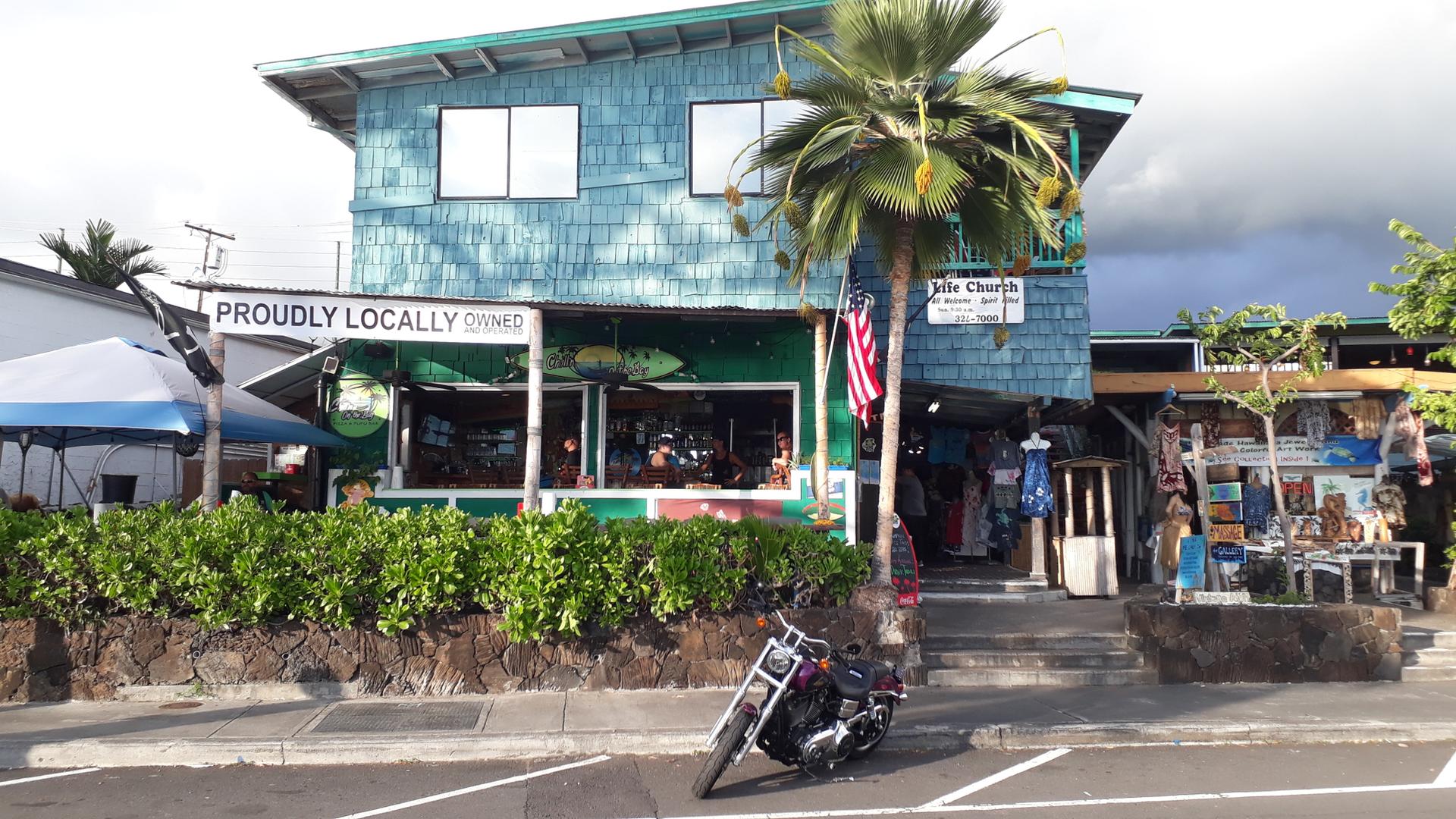 A blue shingled building with shops, a palm tree, and a motorcycle parked outside.
