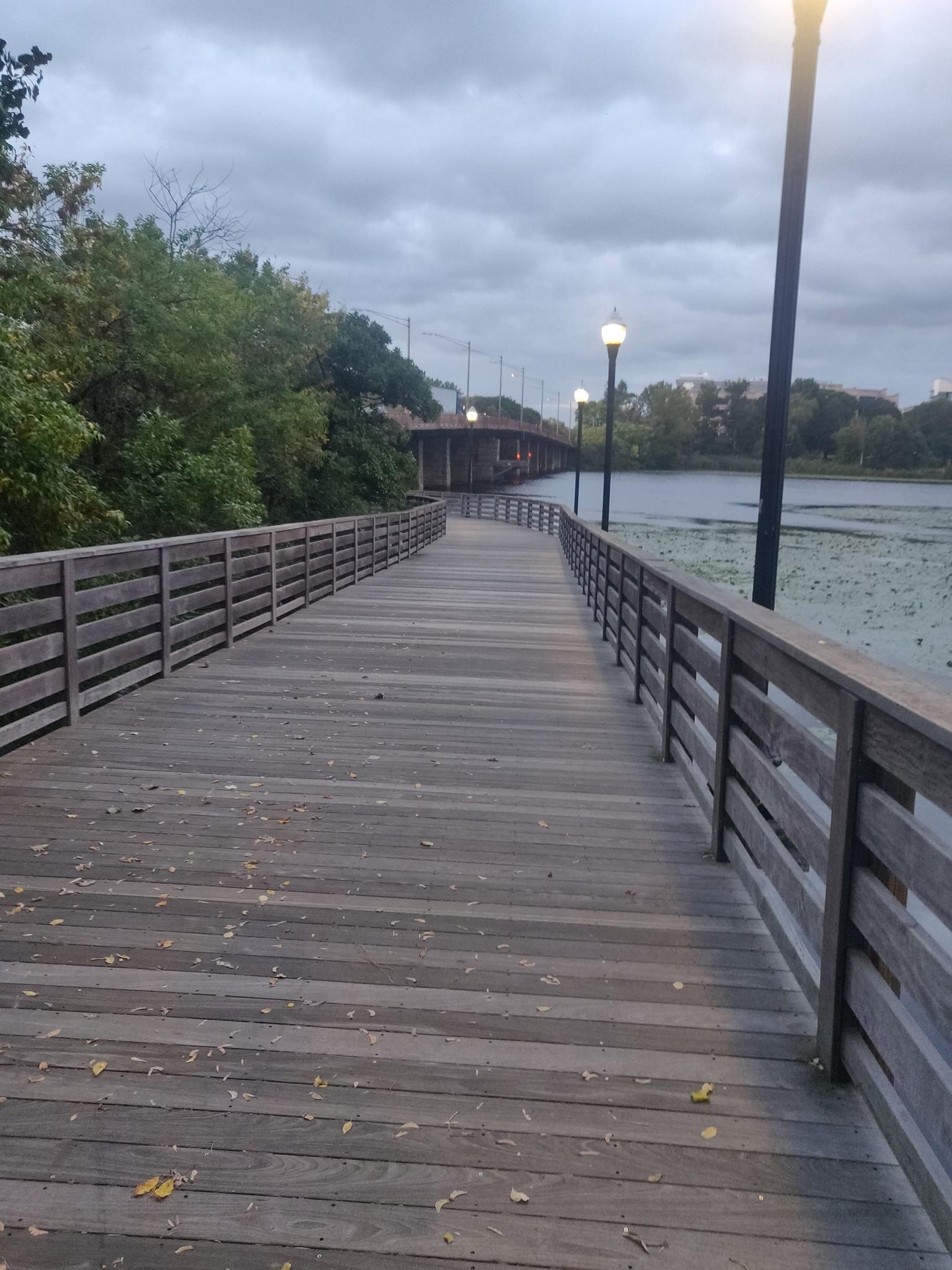 A long wooden boardwalk with railings and streetlights extending over water on a cloudy evening.