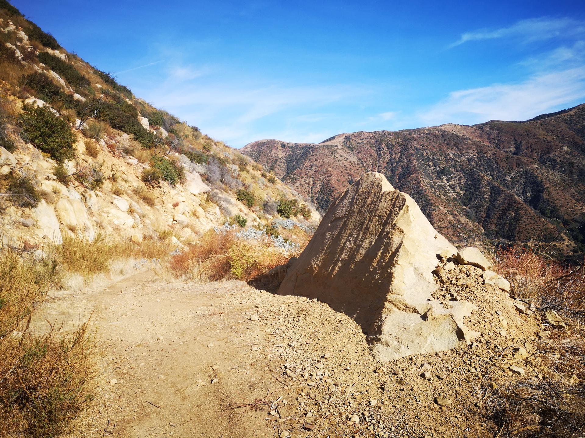A dirt path winds past a large, angular rock on a sunny hillside.