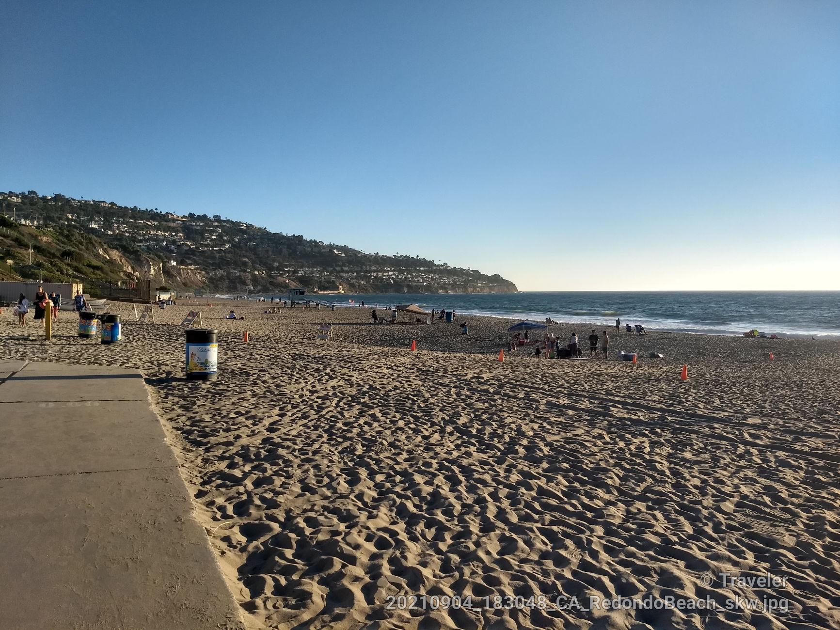 Wide sandy beach with distant people and hills under a clear sky.