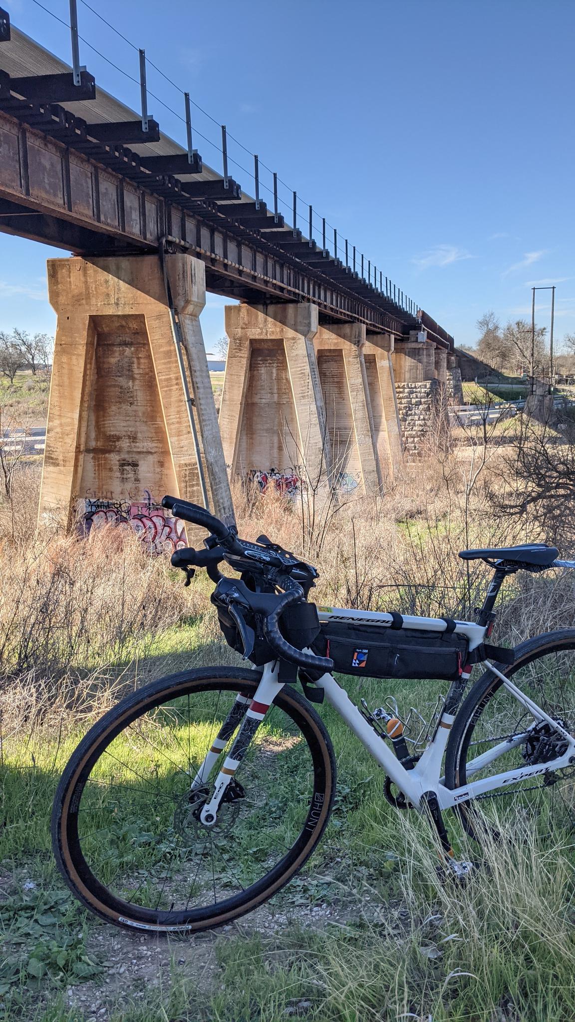 A white bicycle with bags rests under a large concrete and steel bridge.