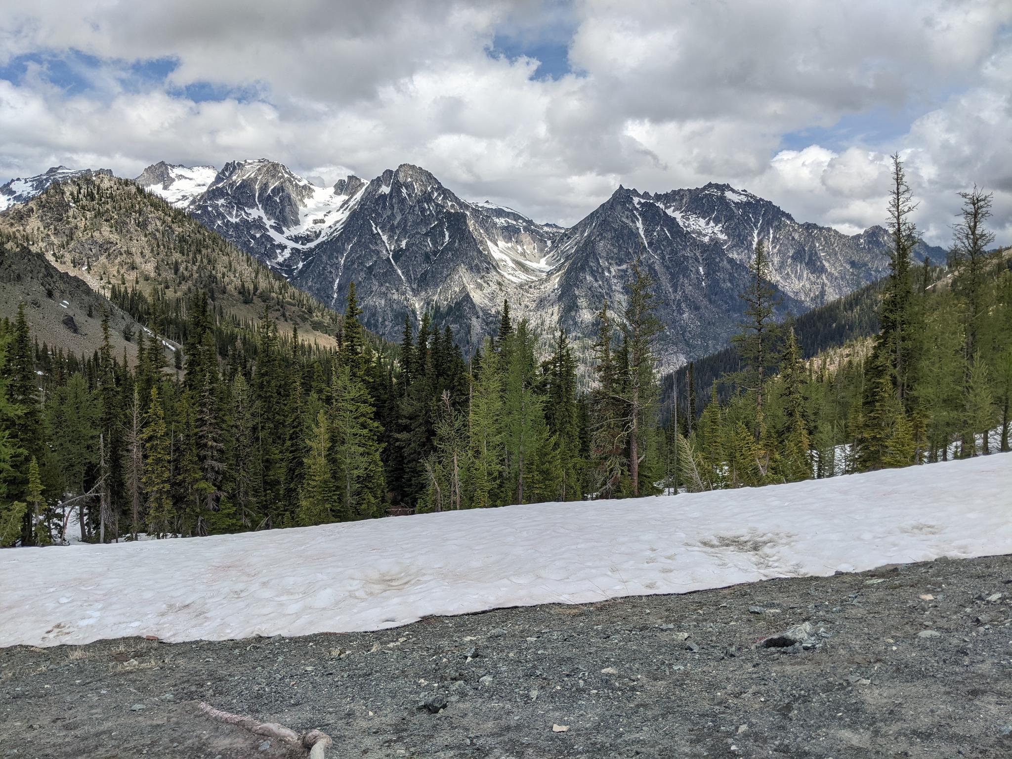 A snow patch in a forest with majestic snow-capped mountains under cloudy sky.