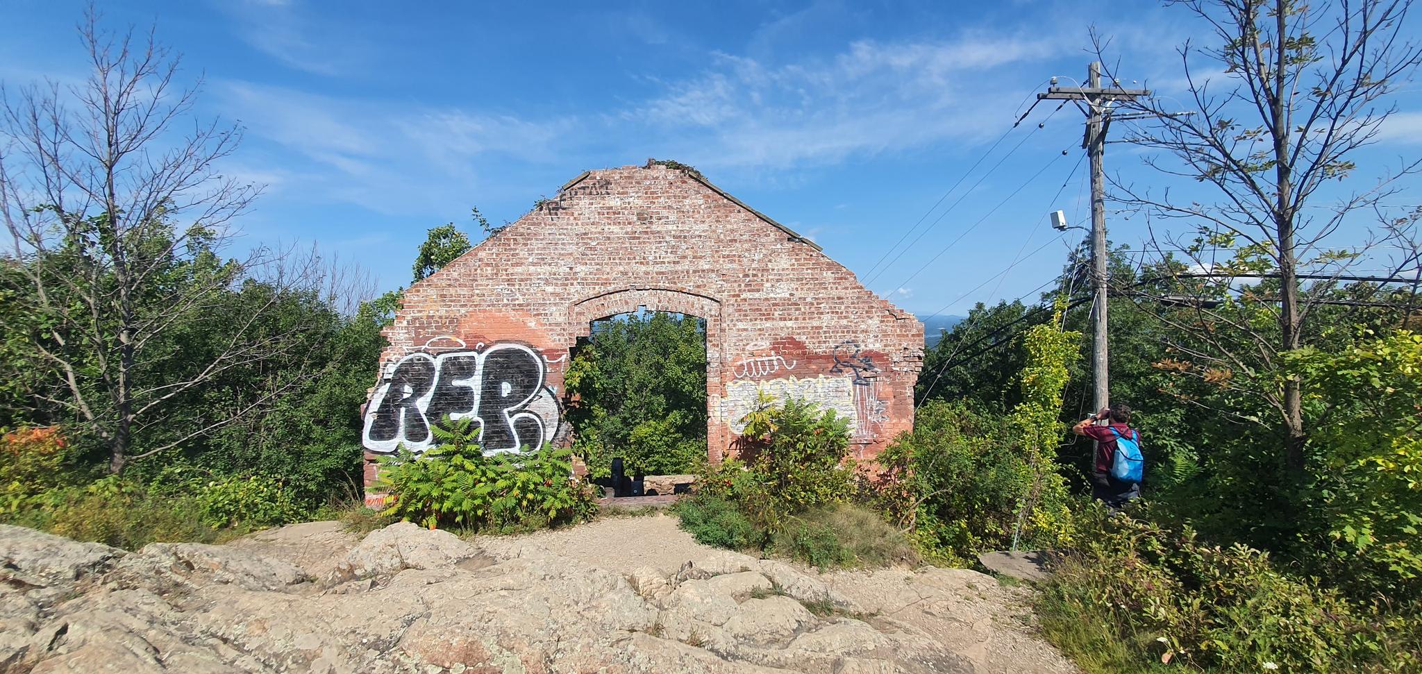 A person stands near a graffiti-covered brick ruin on a sunny day.