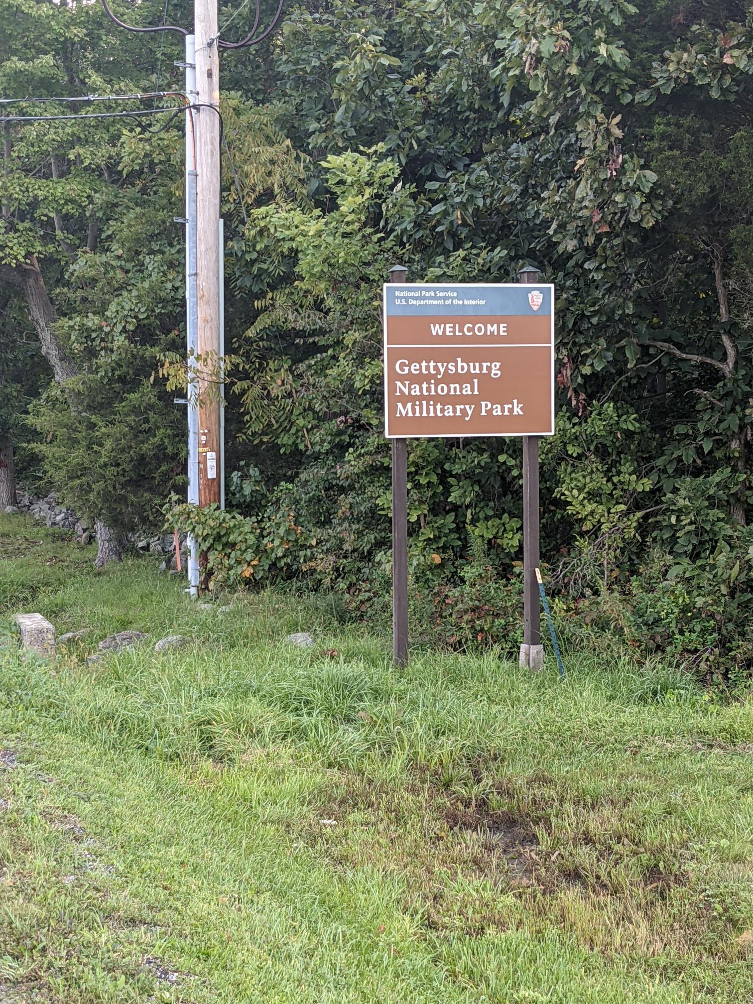 A brown park sign stands amidst dense green foliage and a utility pole.
