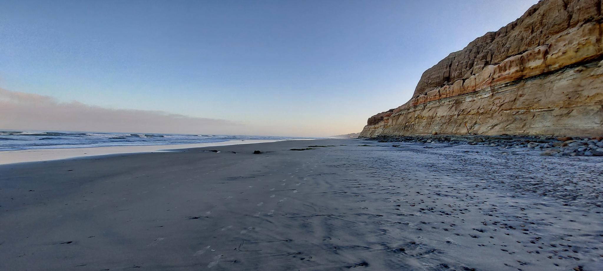 Wide sandy beach next to a tall, layered cliff under a clear sky.