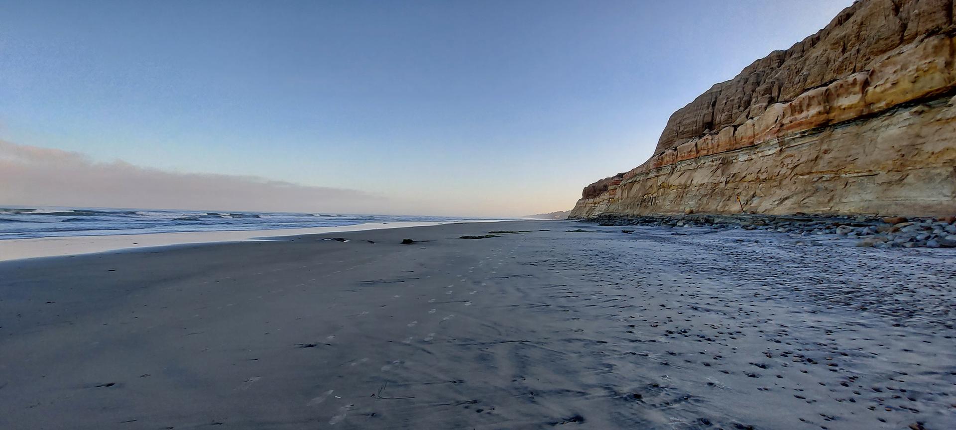 Wide sandy beach next to a tall, layered cliff under a clear sky.
