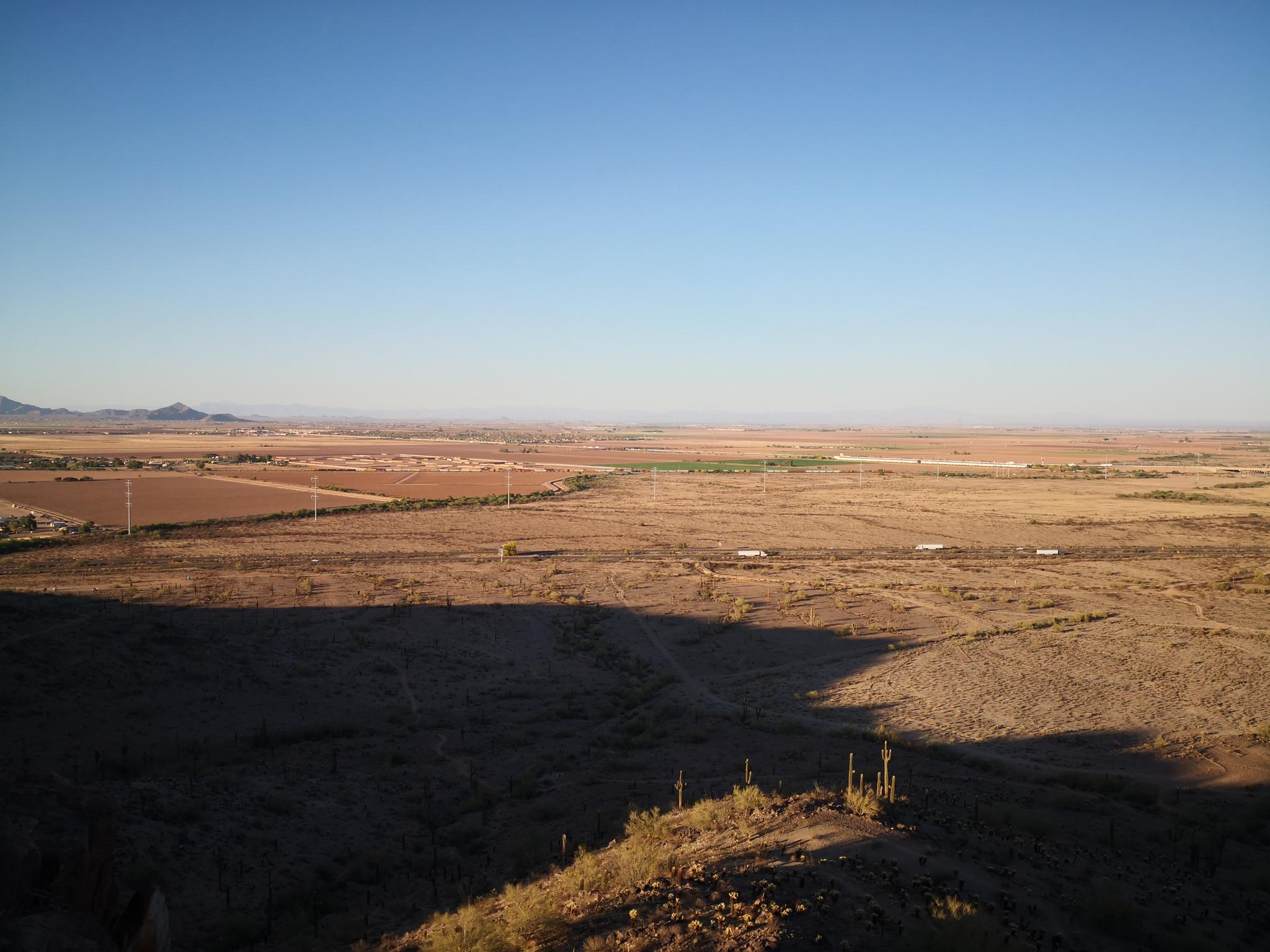 Vast desert landscape with distant mountains, fields, and long shadows under a clear sky.
