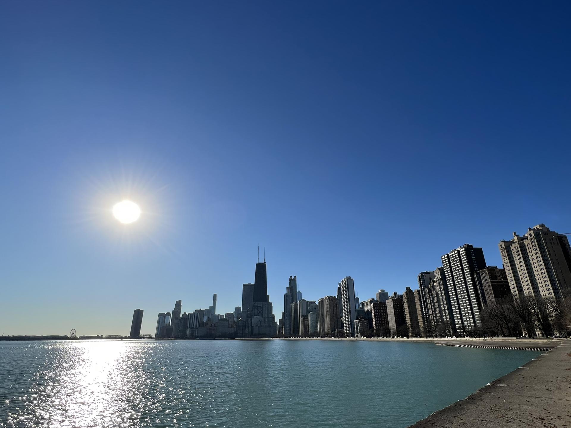 Bright sun over water with a distant city skyline under a clear blue sky.