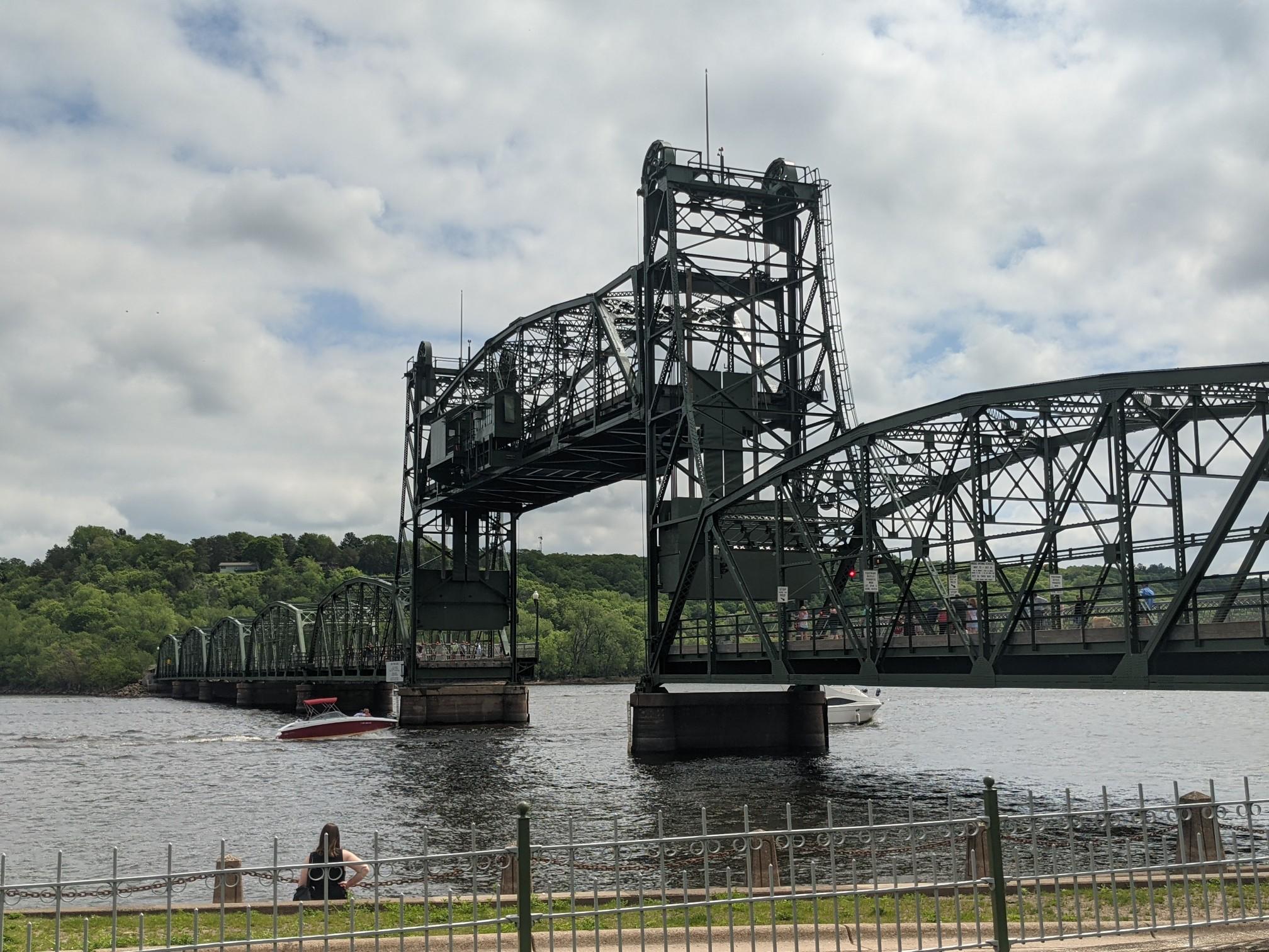 Large green metal bridge over water, person sitting, cloudy day.
