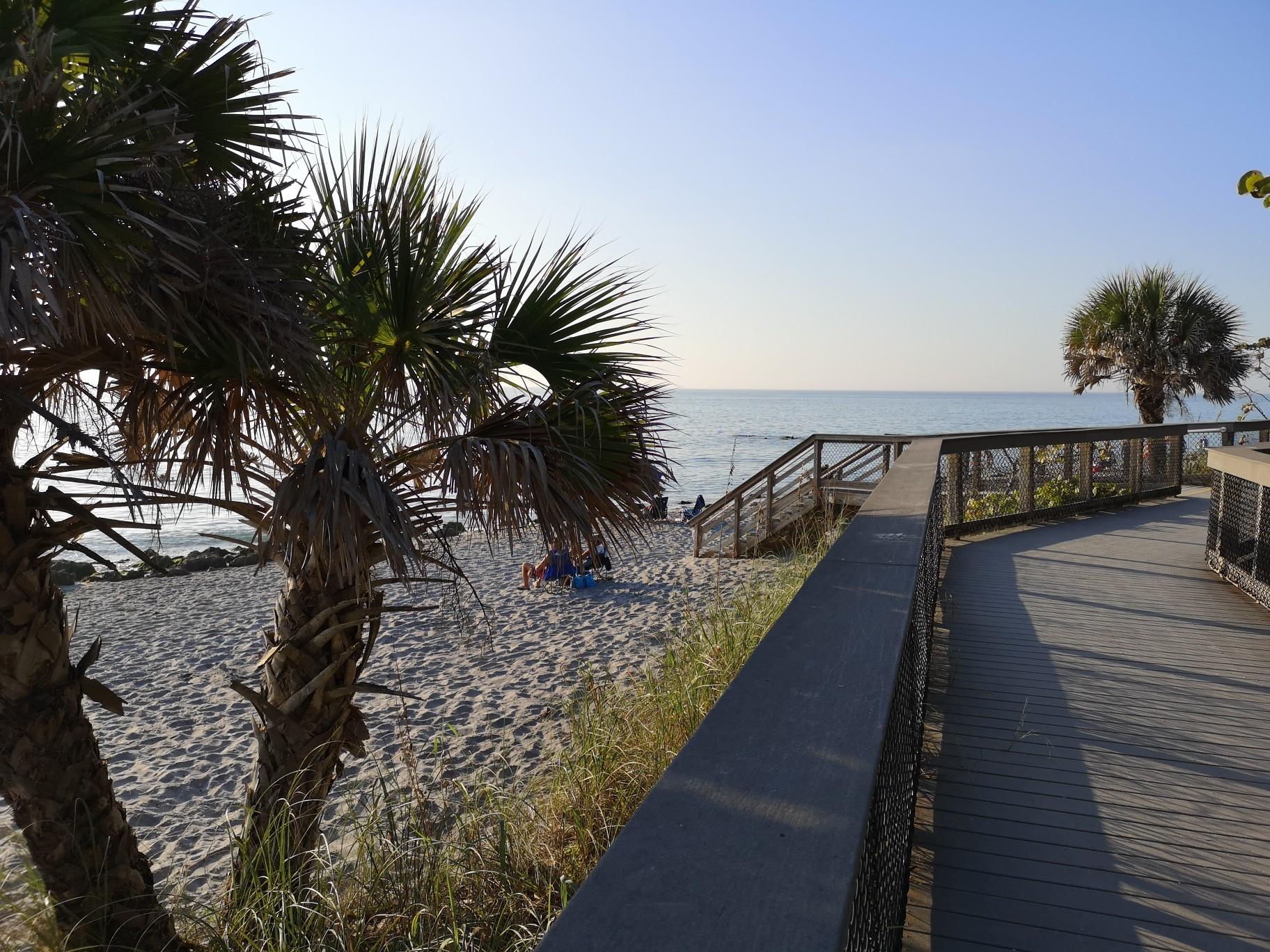 Wooden boardwalk leads to a sunny beach with palm trees and distant people.