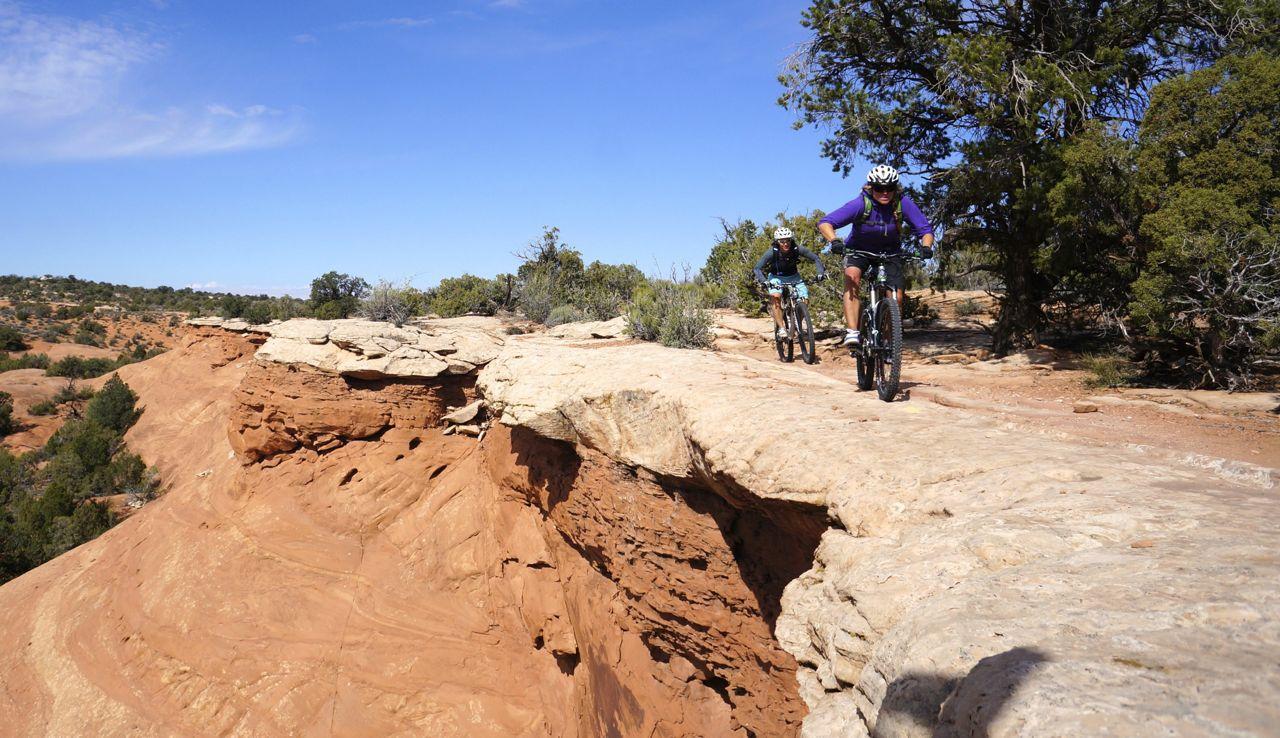 Two mountain bikers ride along a red rock cliff edge under a bright blue sky.