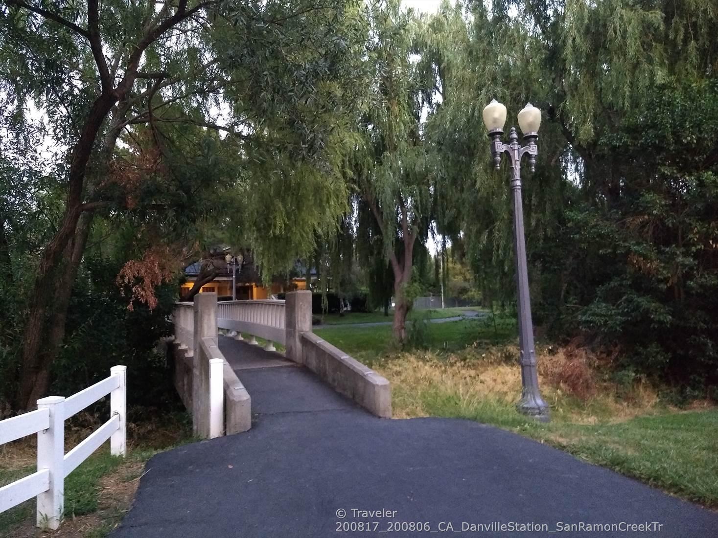Paved path leading to a concrete bridge, surrounded by dense green trees.