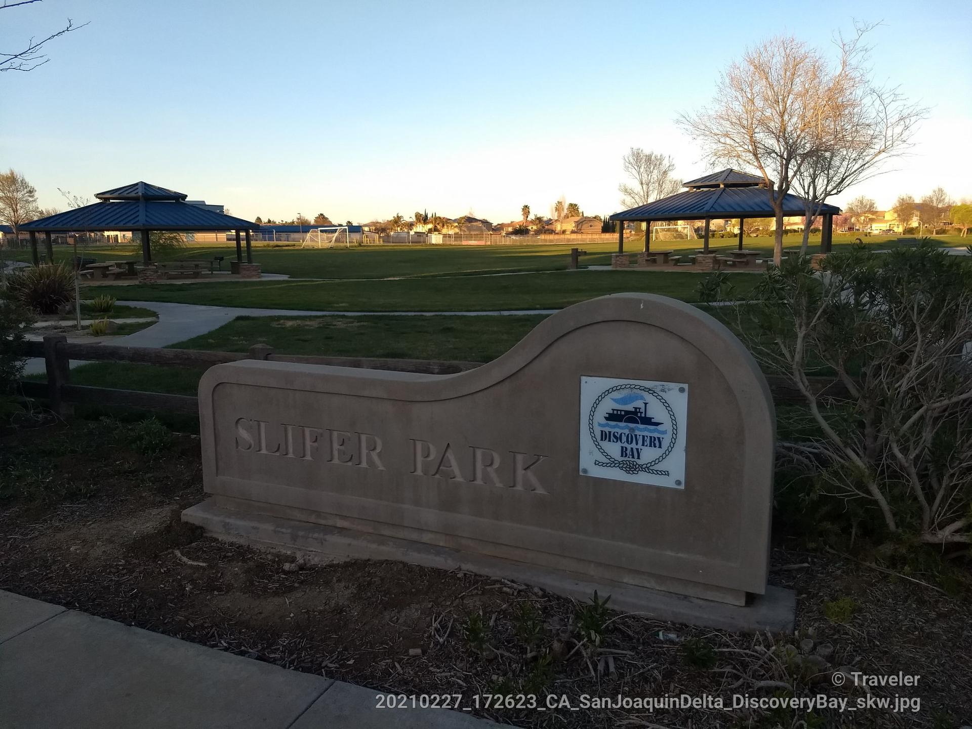 A stone park sign in front of a grassy park with two gazebos.