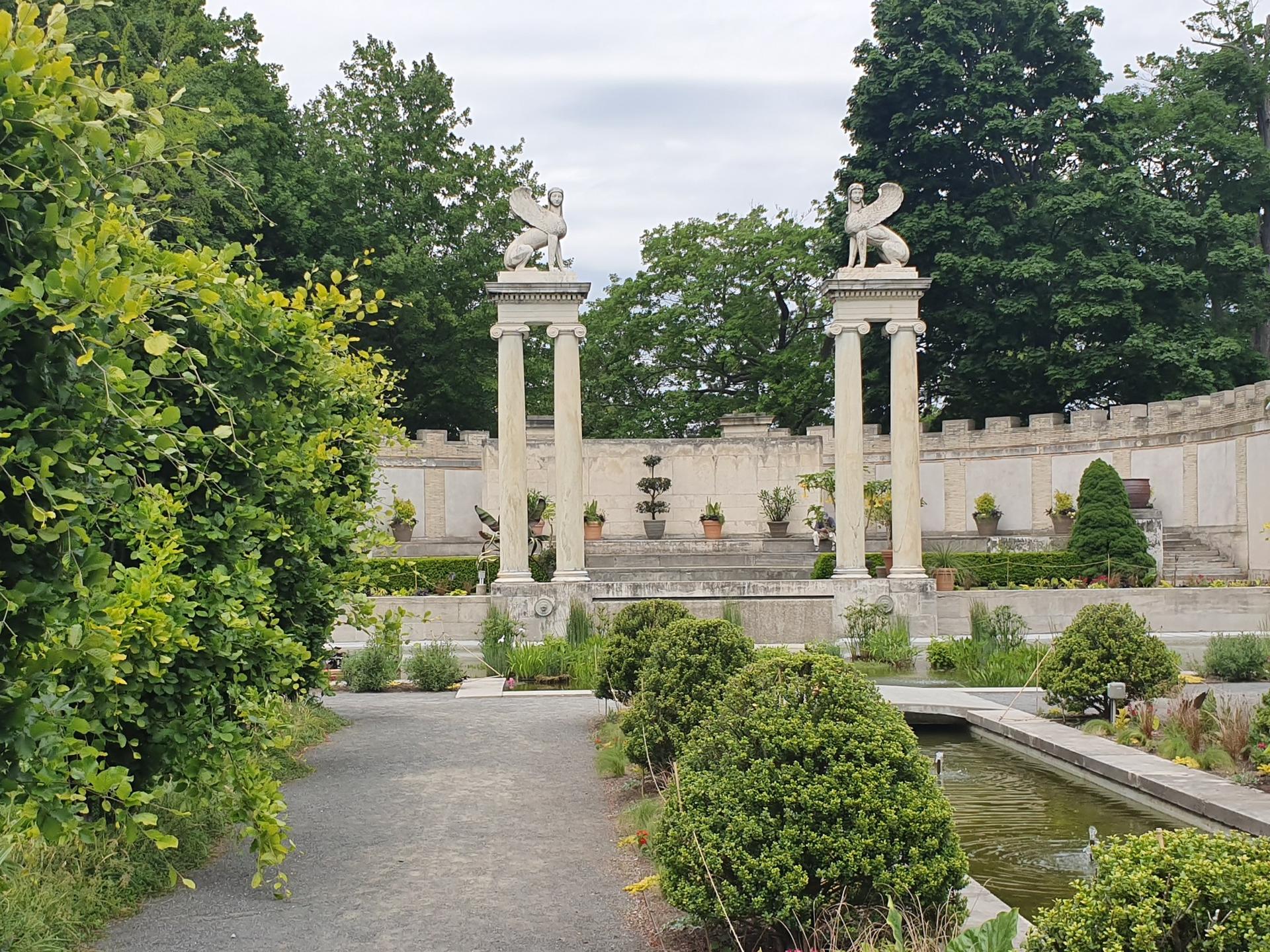 Formal garden with stone columns topped by statues, fountain, lush greenery.
