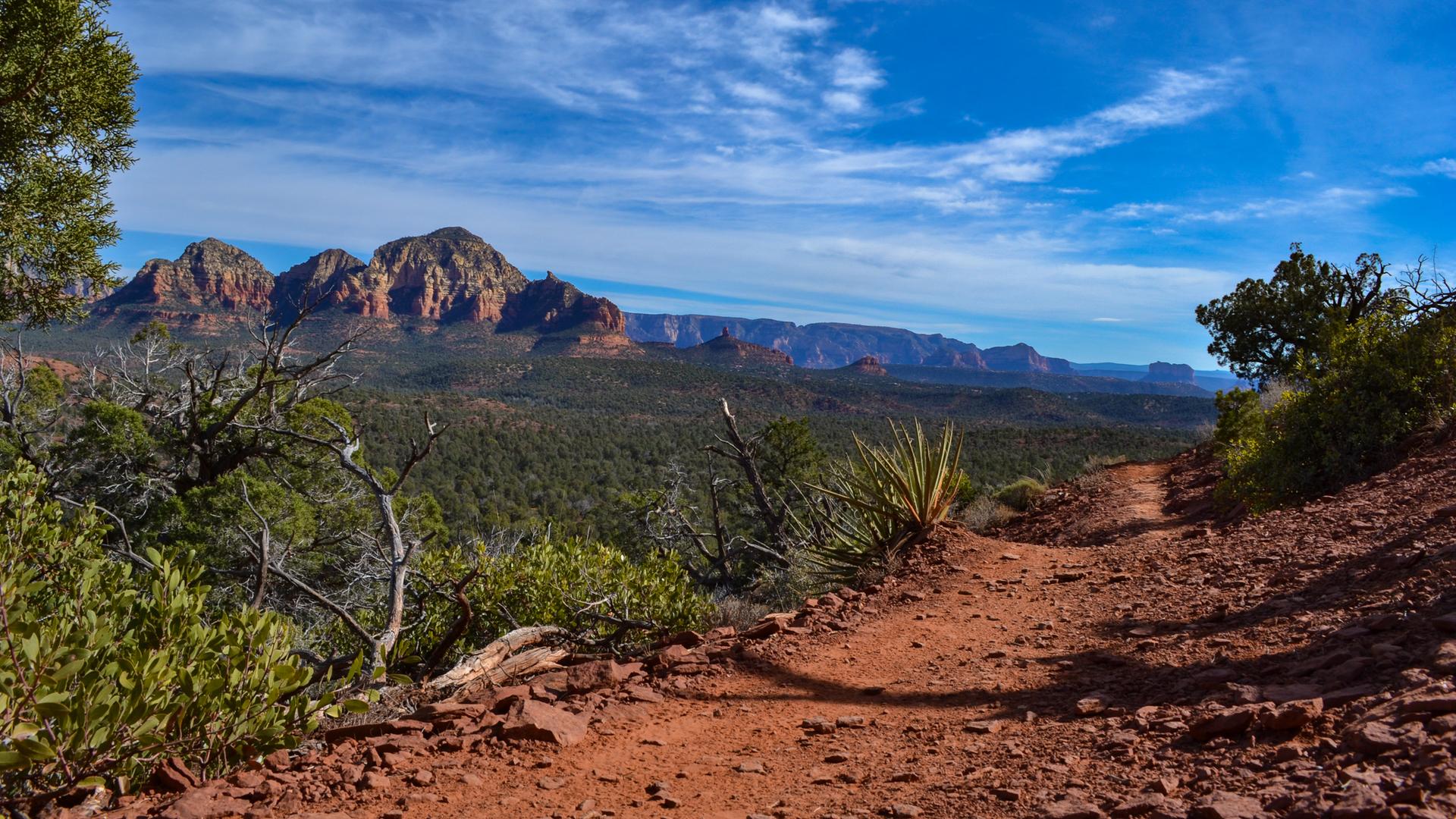 Winding red dirt trail overlooking a vast valley and distant red rock mountains.