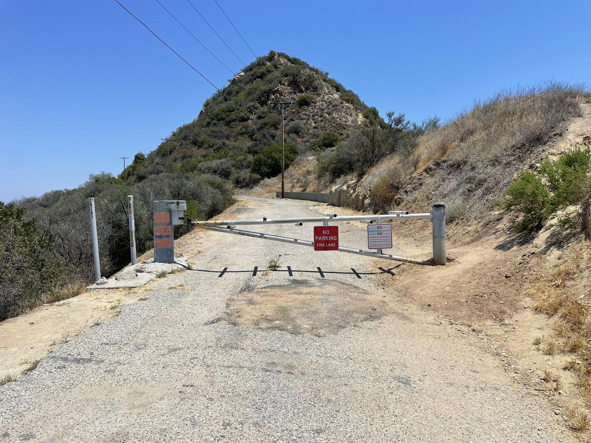 Dirt road blocked by a white gate with red signs on a sunny day.