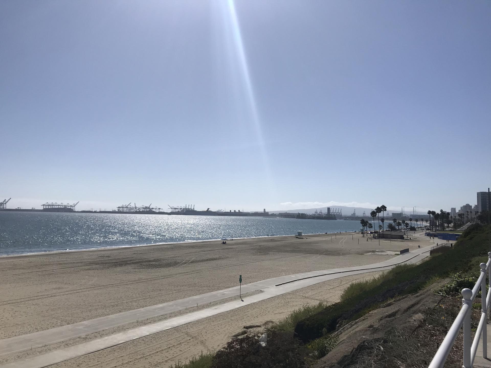 Sunny beach with calm ocean, distant harbor, and bright sky.