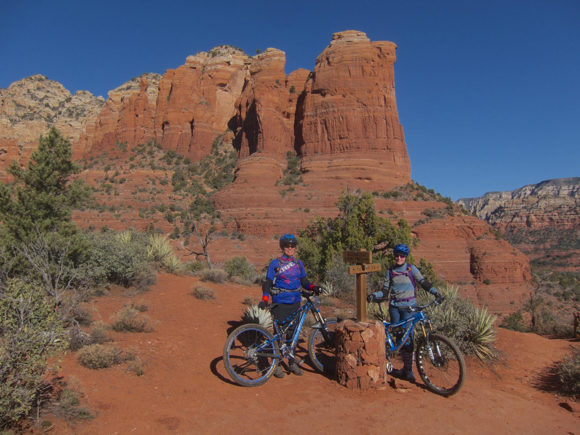 Two mountain bikers stand on a red trail before a massive red rock formation.