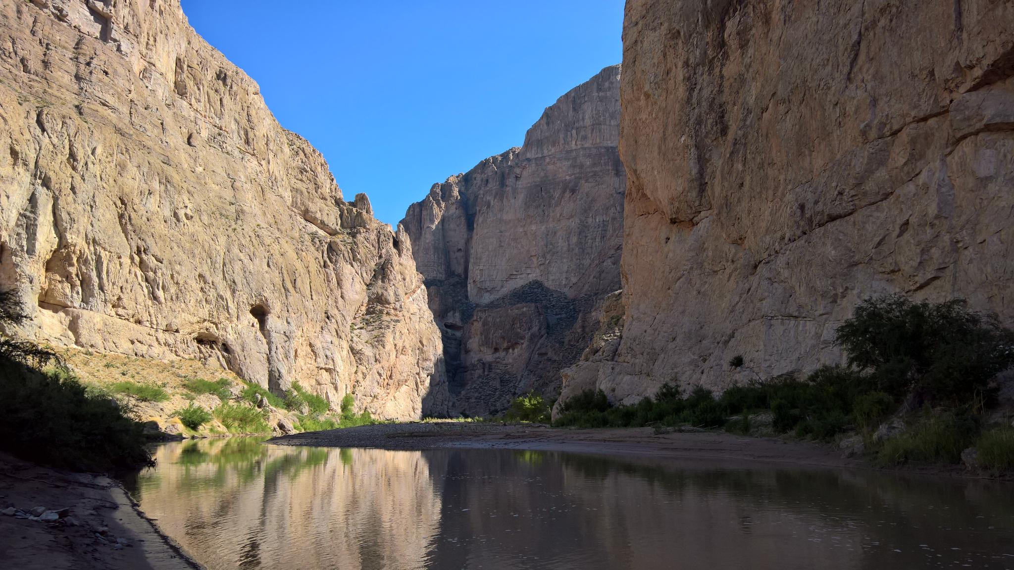A calm river reflects towering canyon walls under a bright blue sky.