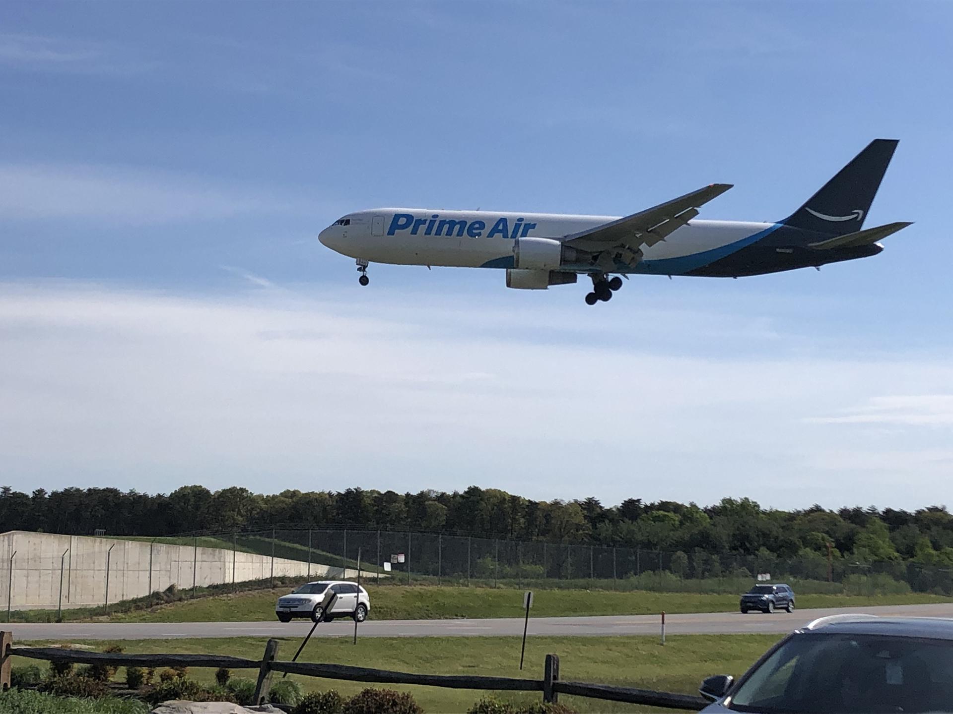 A large cargo plane flies low over cars and trees on a sunny day.