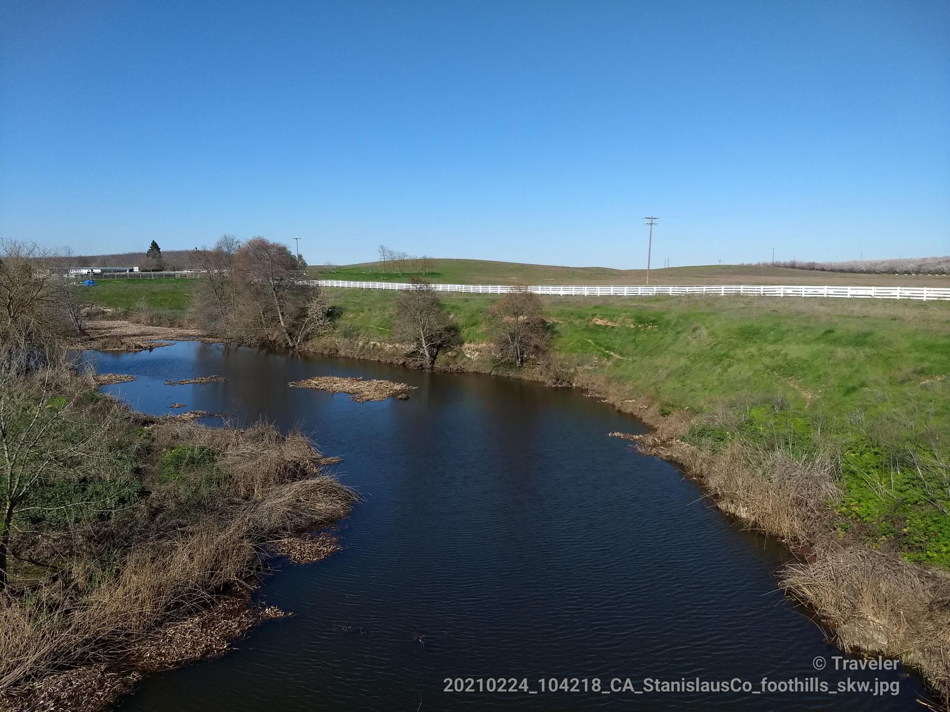 Dark river winding through dry brush towards green hills and a white fence.