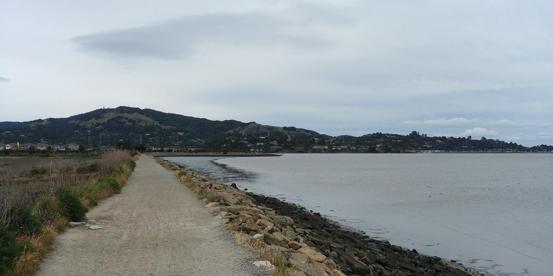 Wide dirt path next to a rocky shoreline and calm water under cloudy sky.