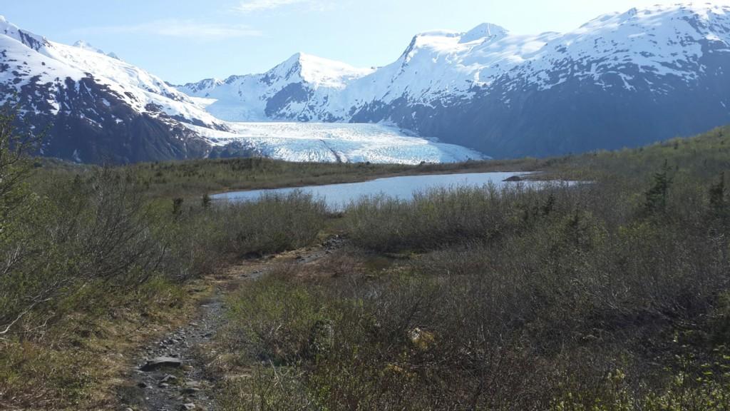 A narrow path leads to a small lake, with a large glacier and snowy mountains.