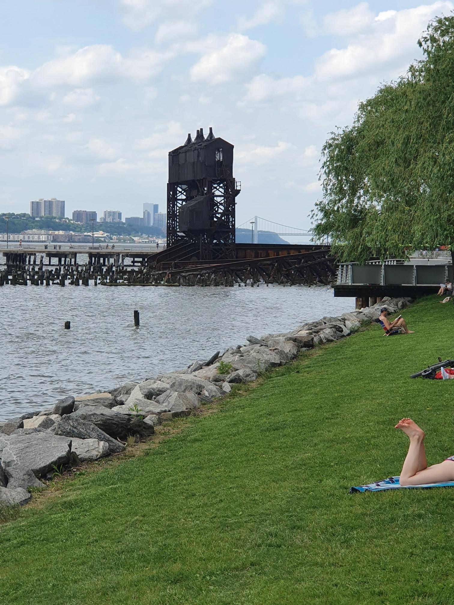 People relaxing on a grassy bank by water with an old industrial structure.