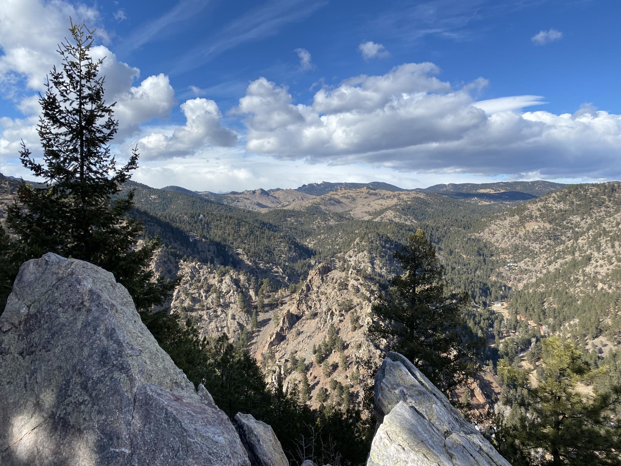 Rocky foreground overlooking a vast, forested mountain landscape under a cloudy sky.