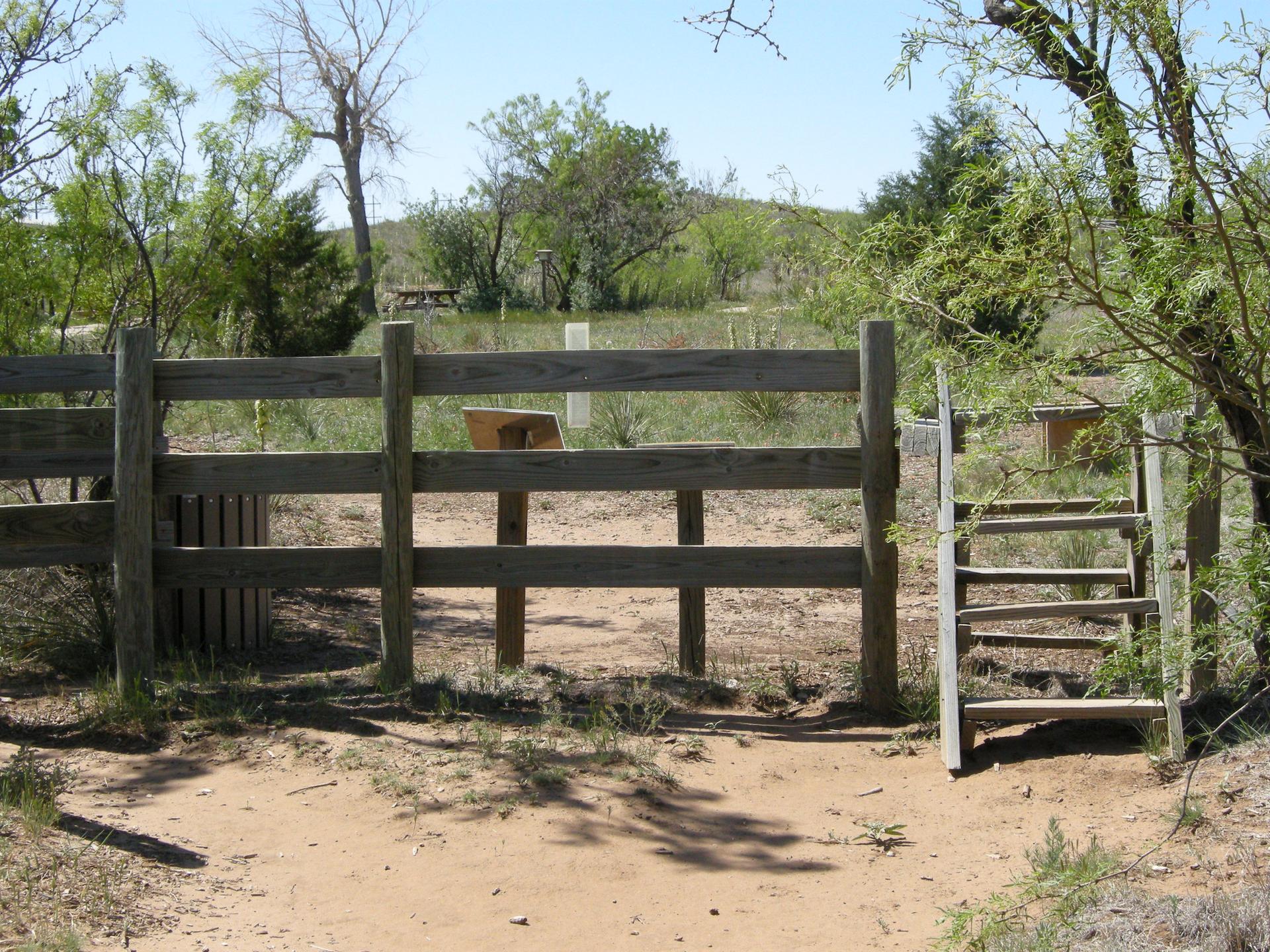 A wooden fence and stile on a dirt path under a bright sky.