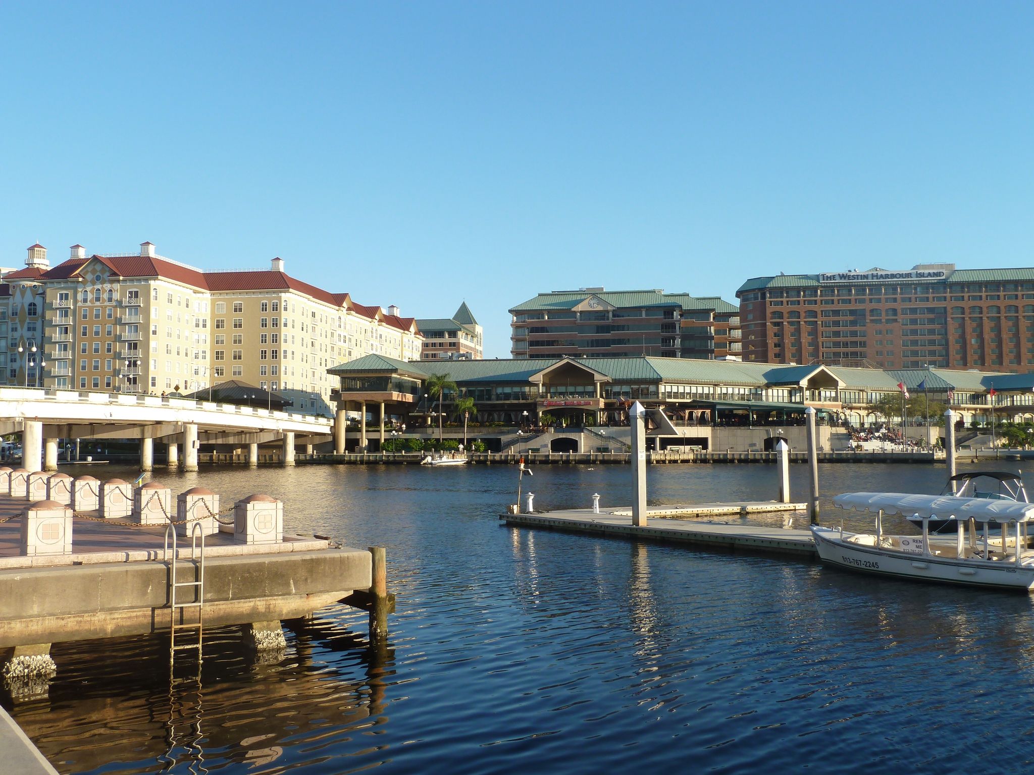Waterfront buildings and docks with a boat on calm water under clear sky. - Tampa Riverwalk