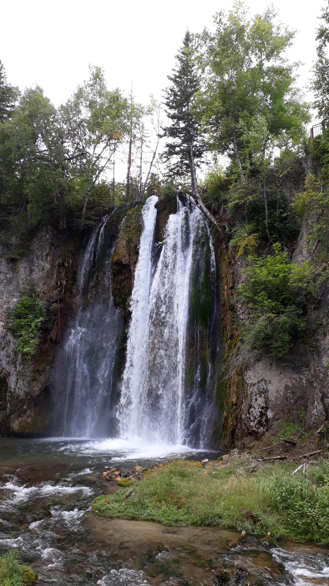 Tall waterfall cascades over rocky cliffs into a stream, surrounded by trees.