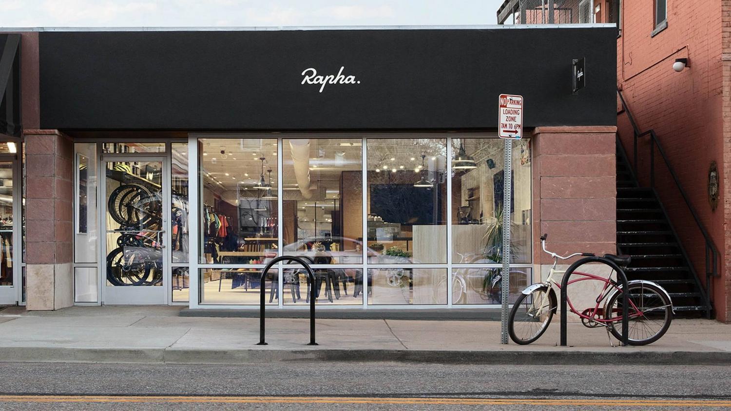 Black storefront with "Rapha" sign, large windows, and a red bicycle outside.