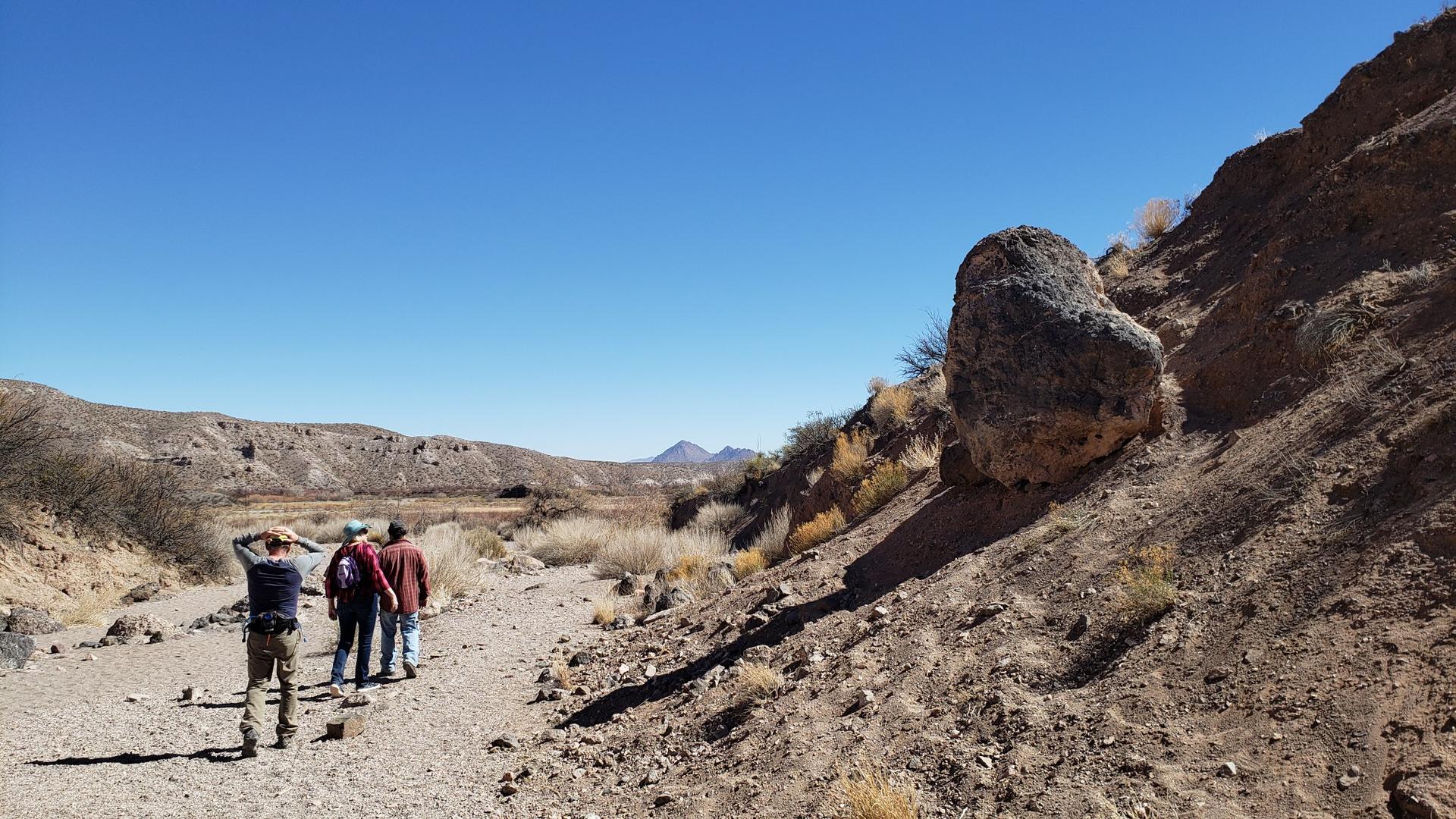 Four people walk along a dry, rocky wash past a large boulder.