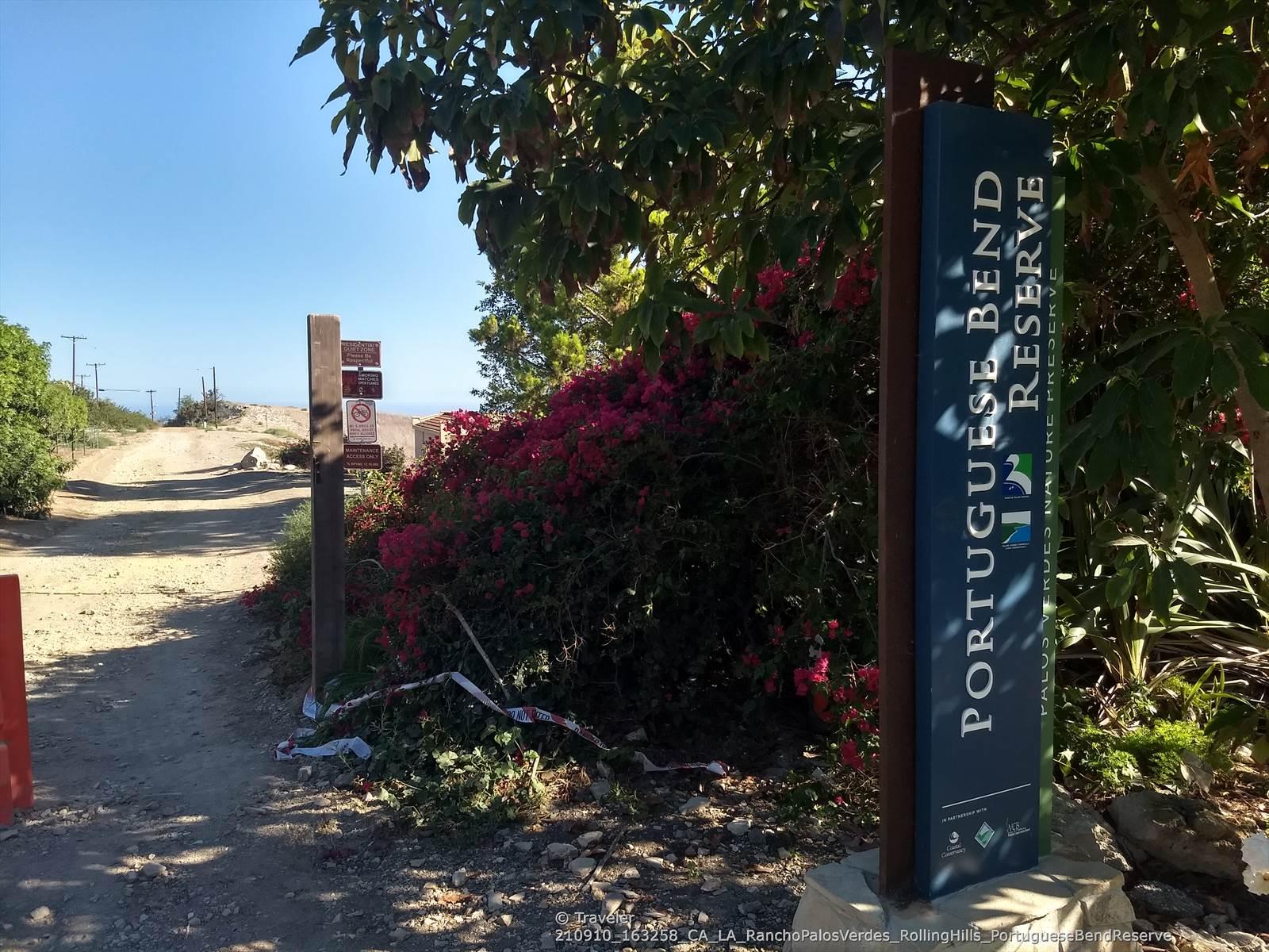 Large sign and lush red flowers beside a dirt road on a sunny day.